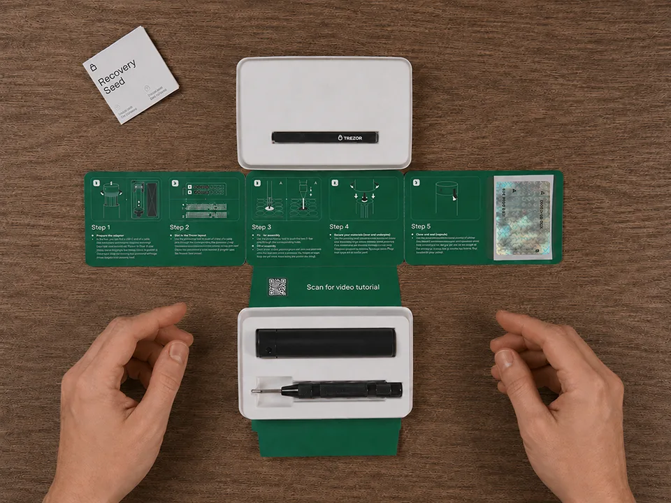 Hands presenting the opened Trezor Keep Metal package with the metal capsule, punch tool, lid, paperclip, and recovery-seed booklet on a wooden desk