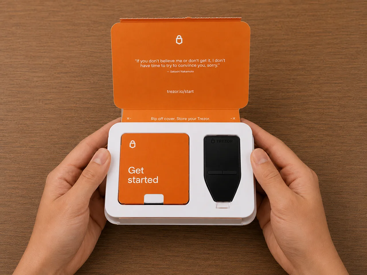 Hands opening the Trezor Safe 3 Bitcoin-Only package with the device and Get started materials visible on a wooden desk